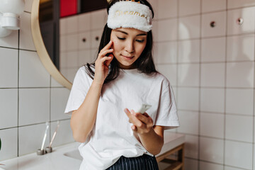 Dark-haired girl in white T-shirt and sleeping mask applies cream to her face and reads its...