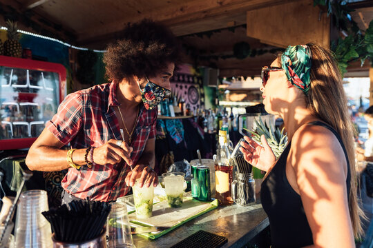 African American barman making refreshing alcoholic cocktail with ice and lime for female client in outdoor bar in summer evening