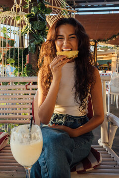 Playful young female making grimace and biting fresh pineapple while chilling in outdoor cafe in summer evening