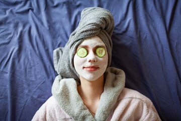 Beautiful young woman with towel wrapped around her head applying clay face mask at the bathroom
