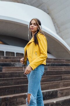 Low angle of young female in trendy jacket and ripped jeans looking at camera while standing on stairs near urban building and listening to music through headphones