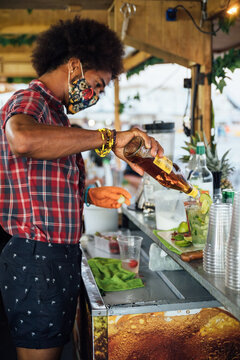 Black male bartender in protective mask adding alcohol into glass with ice and lime while preparing tropical cocktail at counter of beach bar