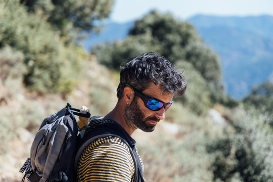 Side view of adult bearded male mountaineer in sunglasses and with backpack standing against blurred background of rocky highland