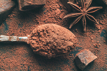 Brown cocoa powder in the spoon, chopped chocolate cubes, top view on dark background