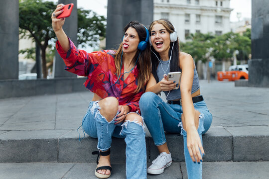 Cheerful hipster female students in trendy outfits taking selfie on smartphone and listening to music with headphones while sitting together on urban street in summer day