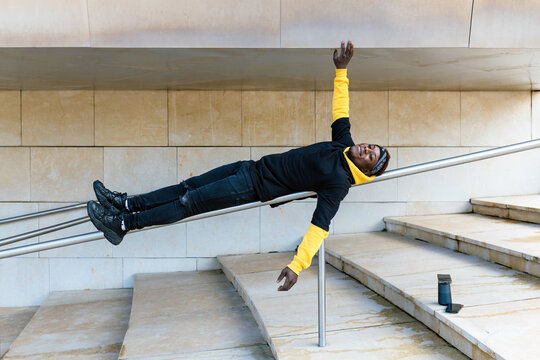 Full body of cheerful young African American hipster male in casual outfit having fun while lying on metal railing near stone stairs with arms outstretched