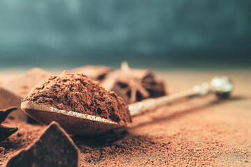 Brown cocoa powder in the spoon and chopped chocolate cubes, on dark background with copy space