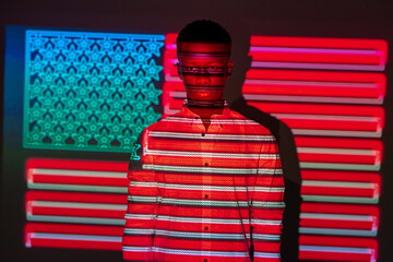Serious African American male illuminated by red and blue neon projection of national American flag standing in dark studio