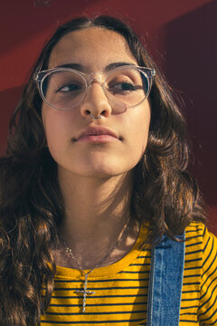 Close up portrait of a thoughtful and introspective teenager girl wearing clear transparent glasses and colorful clothes against red wall with eyes to the side.