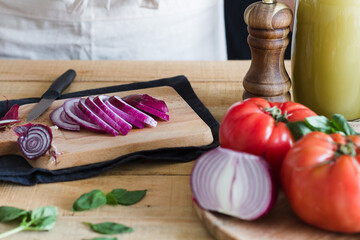 Slices of fresh onion on wooden cutting board while with ingredients for tomato soup in kitchen