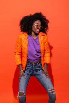 Young black female in purple shirt with denim and orange jacket standing on red background looking down