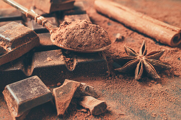Brown cocoa powder in the spoon, chopped chocolate cubes, cinnamon stick and star anise on dark...