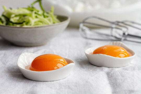 Ceramic bowls with separated raw egg yolks placed on table with fresh greens and mixer during breakfast preparation