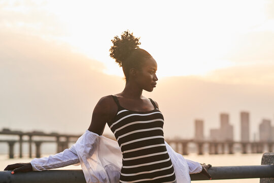 Tranquil pensive young African American female in casual clothes relaxing on urban seafront at sunset time