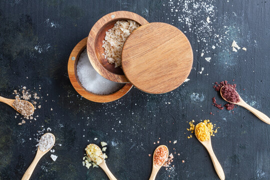 Top view of various types of salt in wooden jars and spoons placed on black table in kitchen