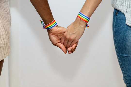 Unrecognizable crop multiracial lesbian couple with rainbow bracelets holding hands on white background