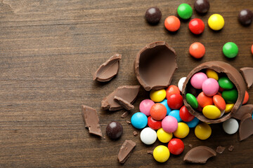 Broken chocolate egg and colorful candies on wooden table, flat lay. Space for text