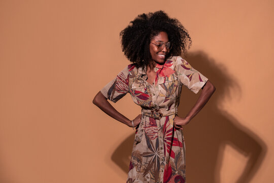 Happy Cuban female in trendy dress and glasses standing with hands on waist on brown background