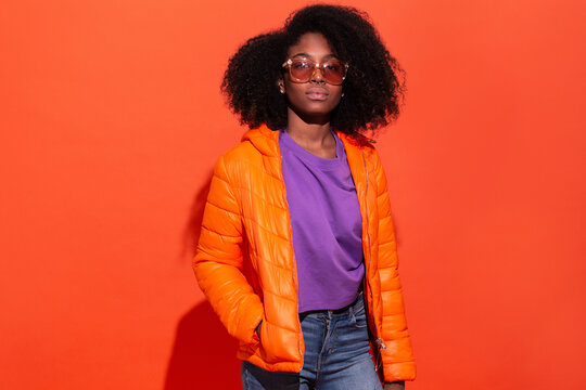 Young black female in purple shirt with denim and orange jacket standing with hands in pockets on red background looking at camera