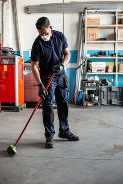 Low angle of man in uniform sweeping floor with broom while working in garage