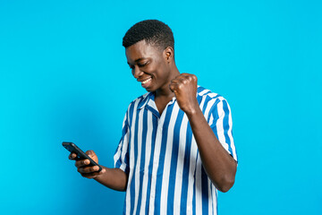 Delighted African American male browsing smartphone and reading good news about business project while standing with clenched fist on vivid blue background in studio