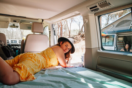 Side view of young female traveler in summer clothes lying in van and chilling on sunny day in woods during vacation while looking at camera