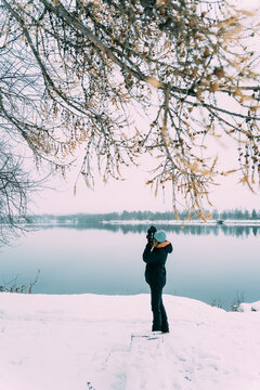 Side view of traveling photographer standing near lake and taking pictures on photo camera while enjoying vacation in winter