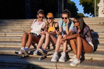 Cheerful young trendy multiracial girlfriends in sunglasses browsing mobile phones while resting together on stone stairs in sunny summer day in city park
