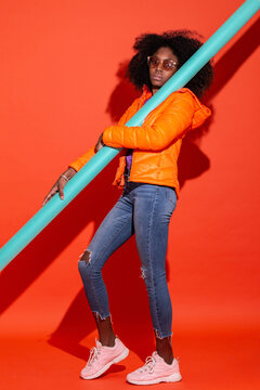 Full body emotionless ethnic woman in colorful jacket holding turquoise pool noodle standing on red background
