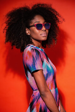 Modern Cuban woman with Afro hair wearing bright dress and sunglasses in red studio background