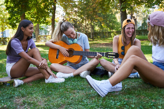 Group of diverse teen girlfriends playing guitar and having fun while sitting together on green grass in park and chilling during summer day