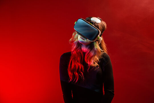 Side view of young unrecognizable blonde woman standing wearing virtual reality headset on colorful red studio background