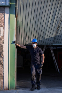 Adult male mechanic in protective glasses and respirator with blue hardhat looking at camera while standing near workshop building on street