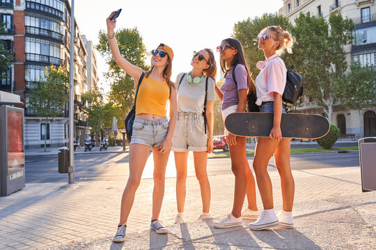 Full body of cheerful trendy young multiracial girlfriends in sunglasses with backpacks and skateboard taking selfie on mobile phone while gathering together on urban square in summer day