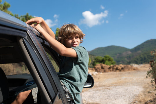 Side view of preteen boy with curly hair sitting in open window of automobile and looking away while enjoying summer adventure in mountainous land