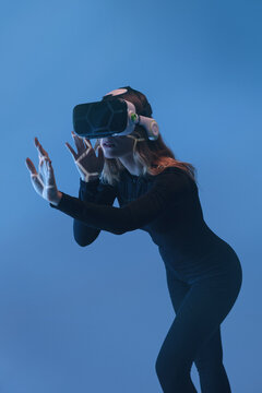 Side view of young unrecognizable blonde woman standing touching air and wearing virtual reality headset on colorful blue studio background