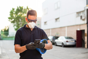 Serious male mechanic in protective glasses and respirator with gloves messaging on mobile phone while standing on street and having break during work in workshop