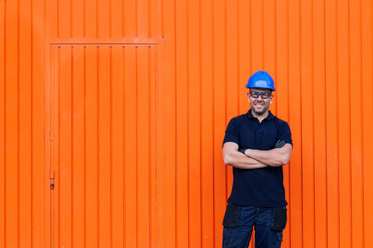 Positive confident adult male workman in blue protective hardhat and eyeglasses smiling friendly while standing against bright orange wall of workshop