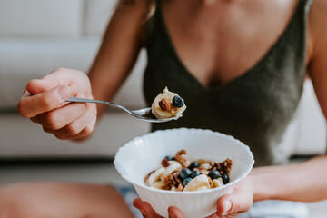 Cropped unrecognizable female with tasty bowl of healthy granola sitting on floor at home and having nourishing breakfast