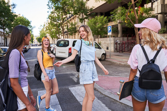 Group of cheerful multiracial teen girls in casual clothes with headphones and skateboard having fun and chatting happily while walking together on city street in summertime