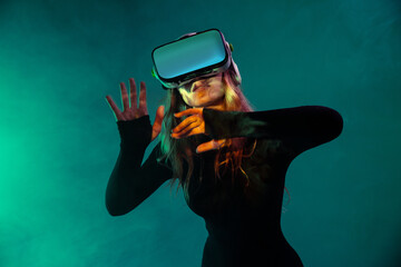 Young unrecognizable blonde woman standing touching air and wearing virtual reality headset on colorful blue neon illumination studio background