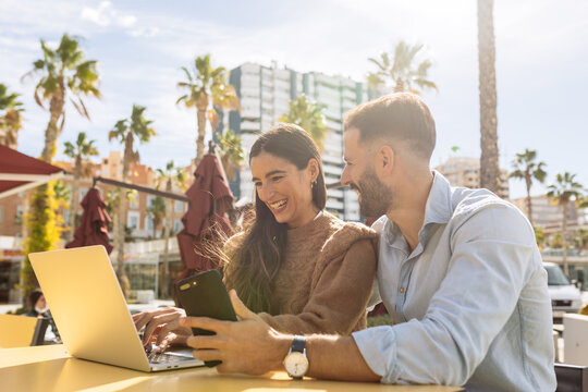 Romantic young ethnic guy in classic outfit hugging and kissing cheek of stylish girlfriend working distantly on laptop in street cafe near sea