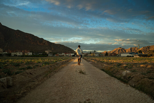 Male tourist lit by sunset sunbeam walking along sandy road in village in highlands - Powered by Adobe