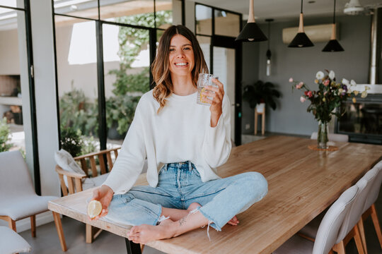 Cheerful female squeezing lemon juice in glass with water while preparing vitamin refreshing drink sitting on top if table at home