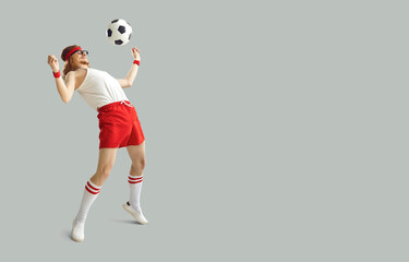 Fototapeta Funny nerdy man having fun with a football. Happy young male sports fan in white and red sportswear kicking away soccer ball on light gray background with free space for advertising text obraz