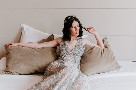 Calm female in sparkling elegant dress resting on comfortable bed with pillows while touching hair and looking away
