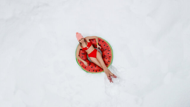 A girl in a red swimsuit lies on an inflatable circle in the shape of a watermelon. Snow, winter, cold. Shooting from above.