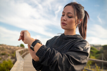 Active young brunette in black sports outfit standing on wooden pathway and checking data on fitness tracker after training in nature