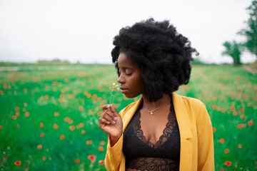 Young curly haired African American female in vivid yellow blazer smelling wildflower while standing in blooming spring field