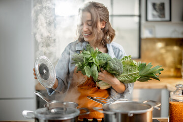 Pretty housewife in apron with fresh vegetables near cooker with boiling pans on the kitchen....